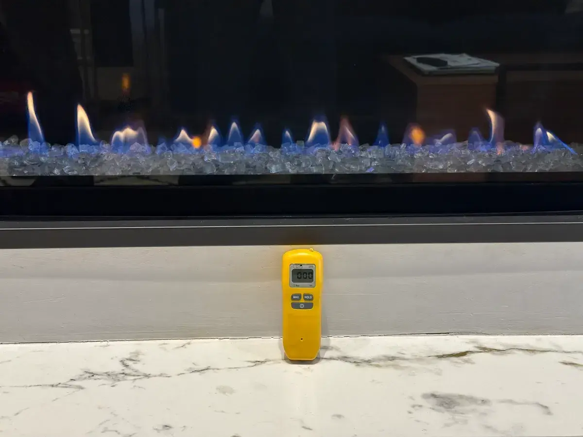 Handheld carbon monoxide meter reading 000 ppm in front of a burning linear gas fireplace