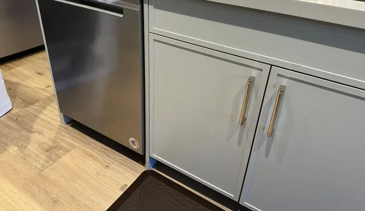 Stainless dishwasher installed in a grey and white kitchen with brass hardware and quartz counters