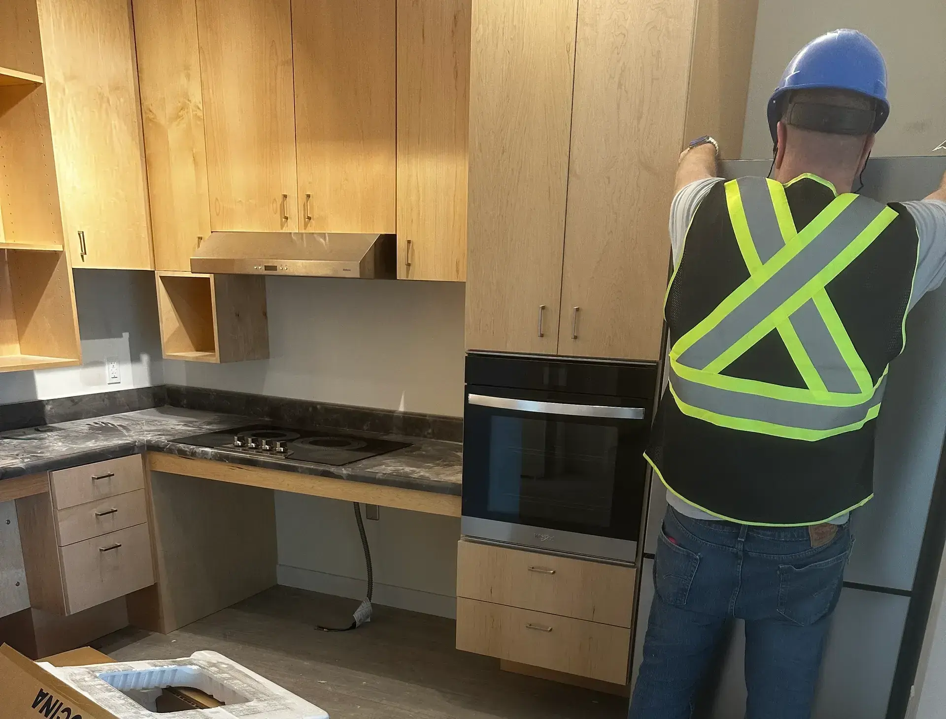Smood Services technician in a hard hat and safety vest setting a built-in appliance beside a new wall oven and cooktop
