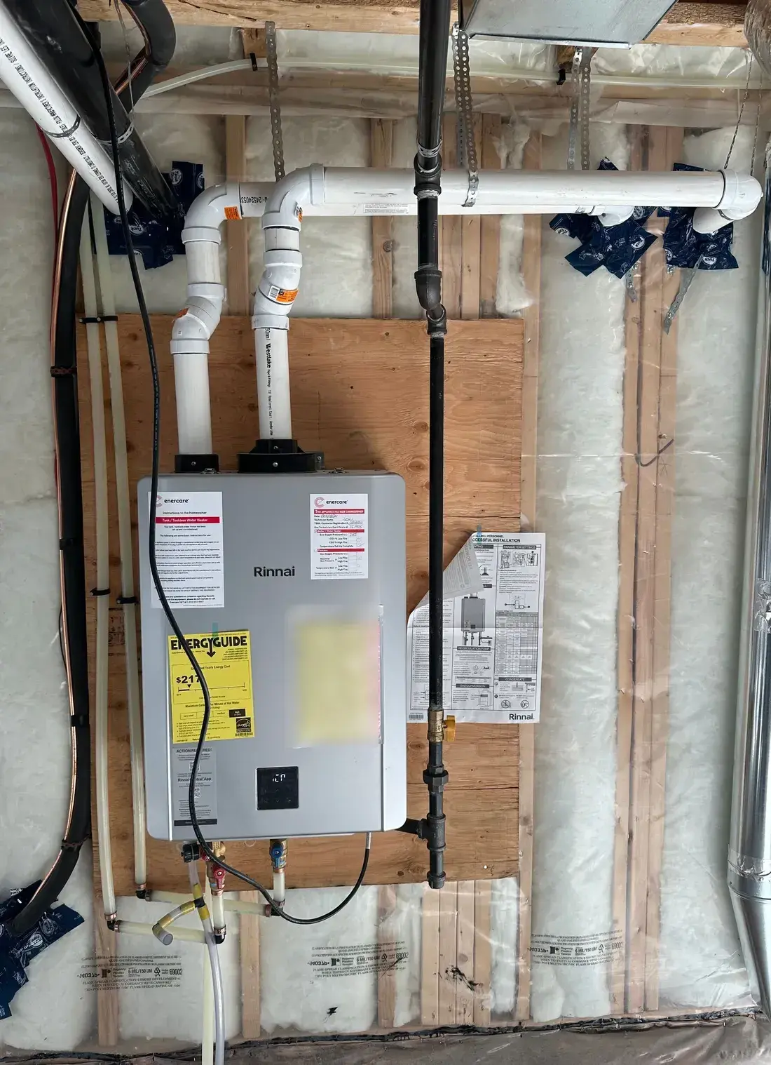 Rinnai tankless water heater installed on a plywood backing board with twin PVC vent runs and a black iron gas line